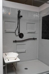 Wall-mounted folding shower seat in a modern shower setting, with a sleek design, gray tiled walls, and a handheld showerhead