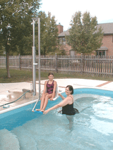 Aquatic Access IGAT-180 pool lift in use, assisting a woman into the pool while another person supports her