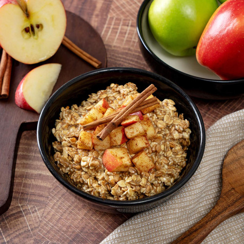 ReadyWise 52 Serving Emergency Food - Prepper Pack Bucket, a bowl of oatmeal topped with apple chunks and cinnamon sticks, surrounded by fresh apples on a wooden table.