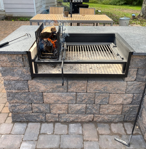 A Uruguayan grill installed into an outdoor stone counter. Wood is actively burning in the vertical brasero on the left, creating embers, and the main grilling grate is positioned over a bed of ash and embers to the right. A grilling tool rests on the counter beside it, with an outdoor dining set visible in the background.