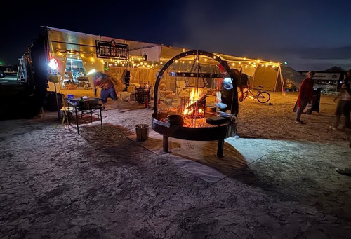 NorCal #5050 Round Catering Grill set up outdoors at night with a roaring central fire basket, overhead arches, and perimeter cooking grates under event lighting.