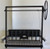 A studio shot of a black steel Argentine grill kit against a plain white background. The kit includes a height-adjustable V-grate suspended by cables, a large flywheel, and a rear brasero basket with a central metal plate featuring a cutout silhouette of a bear and a star.