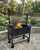 Black steel wood-fired grill with a height-adjustable cooking surface, showing flames and logs in the firebox.