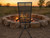 An adjustable Asado Poultry Cross grill cage positioned outdoors in front of a rustic stone fire pit at sunset, showcasing the product base and open-fire roasting setup.