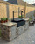 Custom outdoor kitchen featuring a NorCal Ovenworks Santa Maria wood-fired BBQ grill set in a residential backyard with stone masonry and pavers.