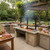 Action shot of a Split Santa Maria Built-In Grill cooking steaks and vegetables simultaneously at different heights, demonstrating dual-zone wood-fired grilling with a live fire in the firebox.