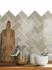 A close-up kitchen counter visual featuring a new backsplash of Layed Greige Herringbone Peel & Stick Composite Wall Tiles (GHT57) with a wooden cutting board, a glass jar of cereal, and bottles.