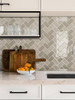 A visualization of a modern kitchen backsplash with Layed Greige Herringbone Peel & Stick Composite Wall Tiles (GHT57) behind cutting boards and a bowl of oranges, with open shelving holding drinking glasses.