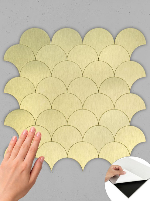 Hand touching the textured surface of a Layed gold metal fan shaped tile sheet, with an inset showing the peel-and-stick adhesive backing.