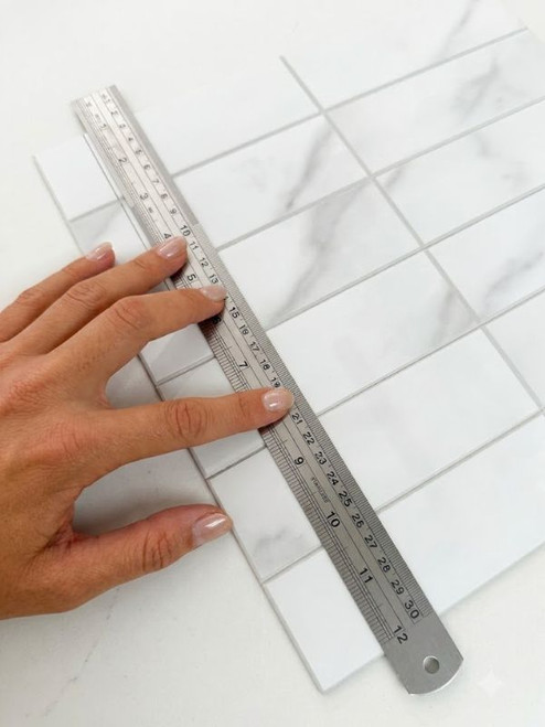 Stainless steel metal ruler held diagonally across white marble peel and stick composite tiles to guide a straight cut.