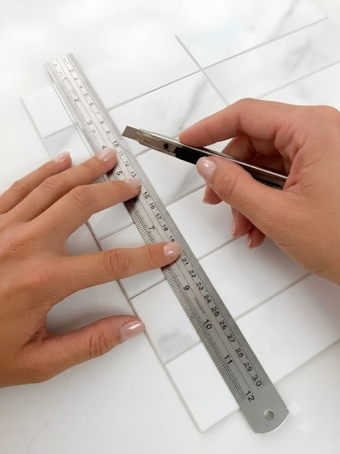 Precision scoring of white marble composite peel-and-stick tile using a utility knife guided by a 30cm stainless steel ruler.