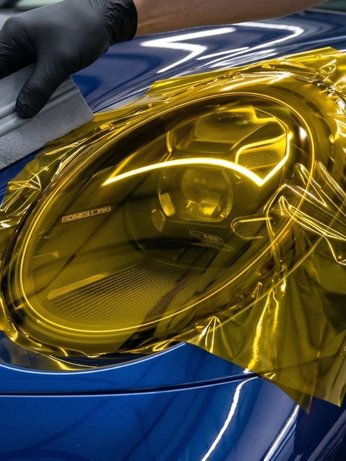 A detailed close-up of a professional auto-detailer in black gloves carefully applying a vibrant, glossy yellow tint film to the sophisticated multi-projector LED headlight of a deep blue Porsche 911 (992), using a gray squeegee tool to smooth the surface and eliminate air bubbles, creating a custom yellow-lens look.