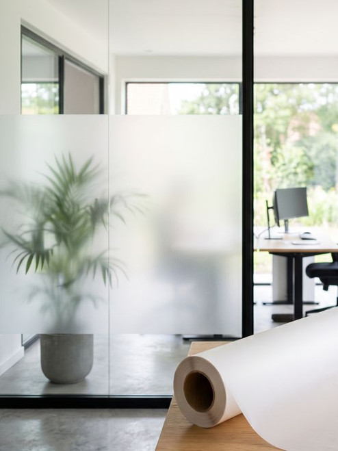 Close-up of a roll of frosted window film on a desk, with a modern office glass partition and a palm plant seen through it, featuring partial frosting for privacy.