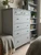 A cozy bedroom corner featuring a tall, multi-drawer dresser beautifully updated with Matte Textured Steel Grey Vinyl Wrap MTSG65, showcasing its smooth, non-reflective painted look alongside a matching bookshelf.
