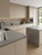 A sleek, modern kitchen featuring handleless flat-panel cabinets wrapped in Matte Textured Taupe Vinyl Wrap MTT212. The soft, earthy taupe cabinetry is beautifully paired with matte grey stone countertops, a black faucet, and warm under-cabinet lighting.
