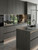 A sophisticated, modern kitchen featuring sleek handleless cabinets and a large central island, all seamlessly wrapped in Lava Grey Oak Wood Vinyl Wrap LGOW17. The dark, straight-grain wood finish is elegantly contrasted by a dark stone countertop, a matte black faucet, and warm under-cabinet LED lighting, with a view of a green garden in the background.