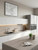 A bright, contemporary kitchen featuring clean white handleless cabinets. The worktops, central island, and matching splashback are flawlessly wrapped in Sonoma Oak Wood Vinyl Wrap SOW33, providing a warm, light woodgrain contrast. The scene is accessorized with a black sink, simple greenery, and a framed map on the wall.