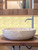 Close-up of a rustic stone bowl basin and modern steel faucet set against a gold metal fan shaped composite tile backsplash.