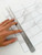 Stainless steel metal ruler held diagonally across white marble peel and stick composite tiles to guide a straight cut.