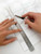 Precision scoring of white marble composite peel-and-stick tile using a utility knife guided by a 30cm stainless steel ruler.