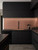 A sleek, moody matte black kitchen showcasing a glowing, warm backsplash completely upgraded with Layed Brushed Metal Copper Vinyl Wrap under modern LED cabinet lighting.