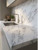A beautifully sunlit kitchen featuring a stainless steel sink set into a continuous countertop and matching backsplash flawlessly wrapped in Layed Volakas White Marble Vinyl Wrap.