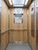 A modern commercial elevator interior featuring wall panels completely transformed with Layed Natural Pine Wood Vinyl Wrap, pairing perfectly with stainless steel fixtures.