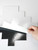 A close-up view of a hand peeling the protective paper backing from a sheet of Layed Greige Herringbone Peel & Stick Composite Wall Tiles (GHT57), demonstrating the simple adhesive installation process.