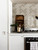 A modern kitchen backsplash visualization showing an installation of Layed Greige Herringbone Peel & Stick Composite Wall Tiles (GHT57) on the wall behind a gas hob and next to white cabinets, with wooden serving boards.
