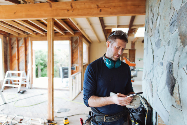 A contractor stands inside a room during renovation, with exposed wood framing in the background and a stone accent wall to his right, as he consults his smartphone.
