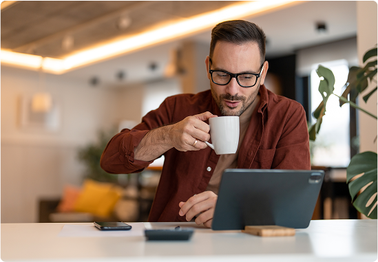 A man working from a modern home office takes a sip from a coffee mug while looking at the screen of a tablet propped up on his white desk.