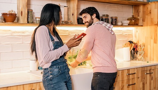 In a warmly lit kitchen with wooden cabinets and under-cabinet lighting, a man at the sink smiles at a woman as they prepare a meal together.