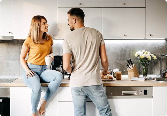 A woman in a yellow shirt sits on the counter of a brightly lit, modern kitchen, holding a coffee mug and talking to a man who is preparing food next to a bouquet of flowers.