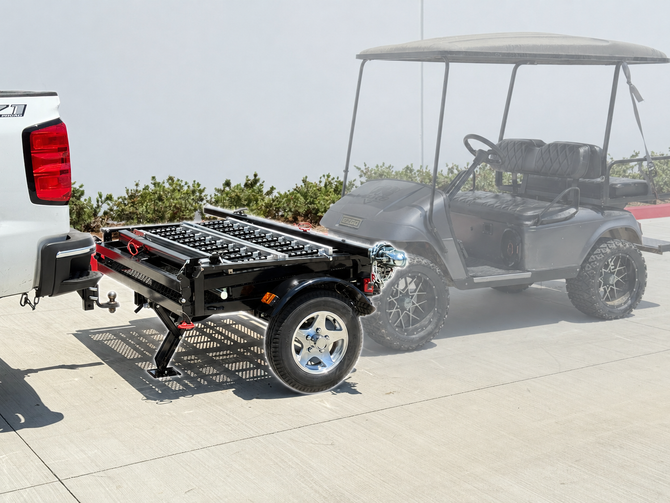 The Compact Caddy folding golf cart and utility trailer hitched to a white pickup truck with a golf cart parked nearby