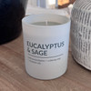 A Eucalyptus & Sage scented soy candle in a minimalist, frosted white glass jar is styled on a wooden table, creating a serene and relaxing home atmosphere.