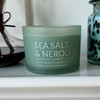 A Sea Salt & Neroli scented soy candle in an elegant, frosted sage green glass jar is styled on a white shelf next to decorative glassware, creating a serene, coastal-inspired scene.