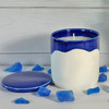 A Summer Days scented soy candle in a beautiful, artisan blue and white ceramic canister is displayed with its lid off, surrounded by decorative blue sea glass to evoke a fresh, coastal feeling.