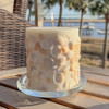A beautiful, hand-crafted inclusion candle covered in real seashells is lit and glowing warmly, sitting on a glass tray on an outdoor table with a scenic coastal view in the background.