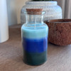 A Volcano Layered Candle in a clear apothecary glass jar with a cork lid, featuring beautiful layers of green, blue, and light blue wax, is styled on a wooden table next to other home decor items.