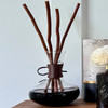 An elegant, glass decanter-style reed diffuser, featuring a vegan leather wrap, is displayed on a wooden shelf, filled with dark fragrance oil and natural willow reeds, creating a sophisticated home decor statement.