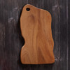 A natural edge walnut wood cutting board with a unique organic shape and a finger grip cutout, displayed on a dark, textured background.