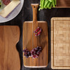 A teak paddle board with grapes and cheese, showcasing its rustic charm and extended handle, against a dark wooden surface.