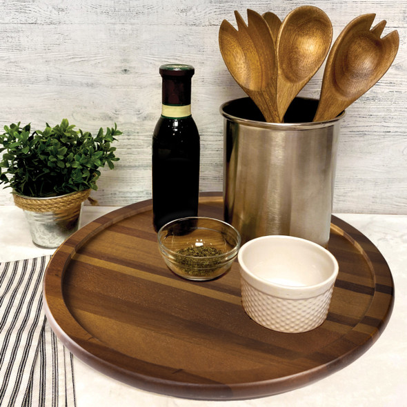 A large, round Acacia Lazy Susan is elegantly styled as a tabletop centerpiece, holding a bottle of wine, a utensil crock, a small plant, and condiment bowls, showcasing its use for organization and serving.