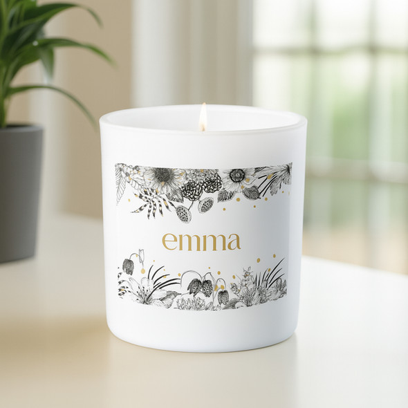 Personalized white candle with the name 'emma' in gold lettering, featuring a black and gold floral design, lit and sitting on a light-colored surface near a window with soft natural light and a potted plant in the background.