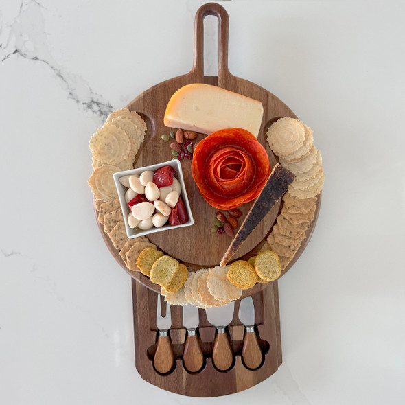 A stylish, round acacia wood cheeseboard is elegantly arranged with assorted cheeses, crackers, nuts, and a salami rose on a marble countertop, with its slide-out drawer open to reveal a set of cheese knives.