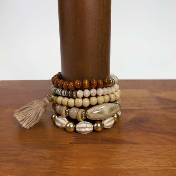 A stylish, five-strand Boho Mixed Bracelet Stack in neutral tones of tan, brown, and off-white is displayed on a wooden stand, showcasing its blend of wood beads, stones, and a tassel accent. A stylish, five-strand Boho Mixed Bracelet Stack in neutral tones of tan, brown, and off-white is displayed on a wooden stand, showcasing its blend of wood beads, stones, and a tassel accent.