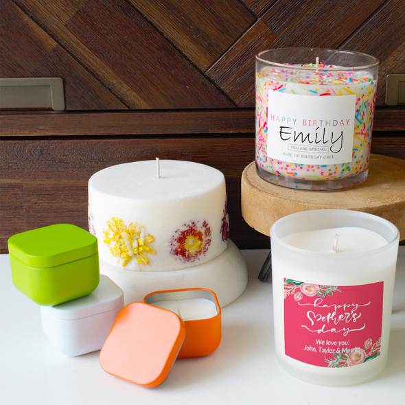 The large Sweet Blooms Inclusion candle is shown in a lifestyle setting alongside a set of smaller, colorful square candle tins, highlighting it as a statement piece in a larger collection.