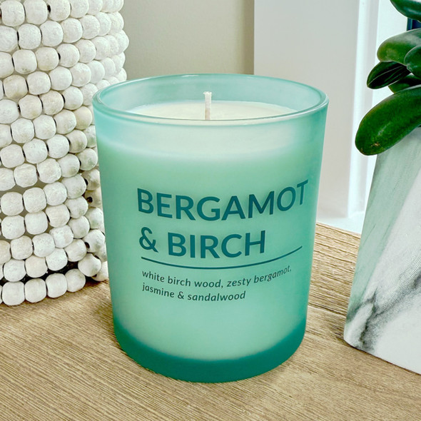A Bergamot & Birch scented soy candle in a sophisticated, frosted seafoam green glass jar is styled on a wooden surface next to a decorative white beaded vase and a houseplant.
