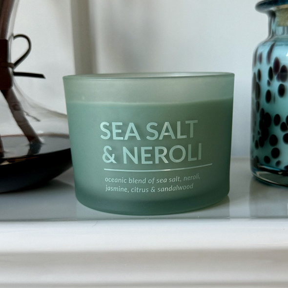A Sea Salt & Neroli scented soy candle in an elegant, frosted sage green glass jar is styled on a white shelf next to decorative glassware, creating a serene, coastal-inspired scene. A Sea Salt & Neroli scented soy candle in an elegant, frosted sage green glass jar is styled on a white shelf next to decorative glassware, creating a serene, coastal-inspired scene.