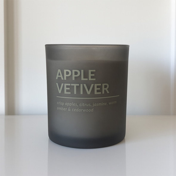 An Apple Vetiver scented soy candle in a sophisticated, matte black frosted glass jar is displayed in a clean, modern setting.